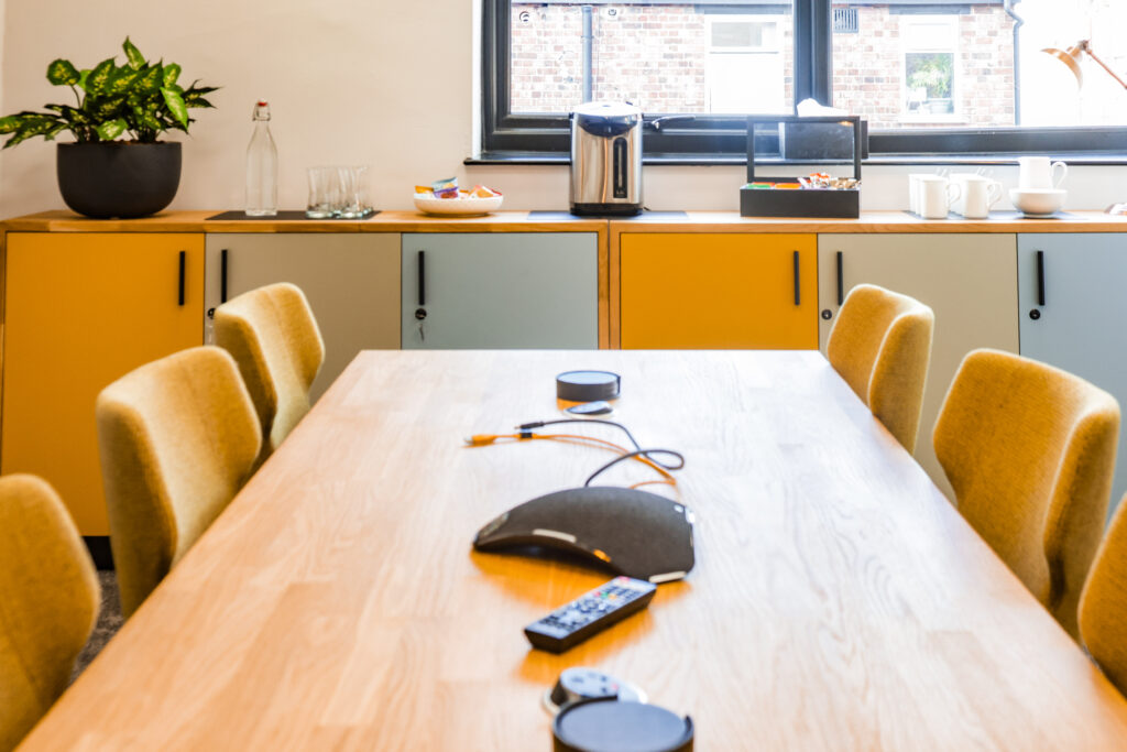 Shared desk space with power access. Six chairs around a large table with tea and coffee facilities nearby.